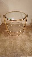 Full view of the round display table showing brass finish metal frame with glass top and glass lower shelf on carpeted floor.
