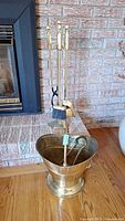 Overall view of the fireplace tools set on stand with brass coal bucket in front on wooden floor near brick fireplace.