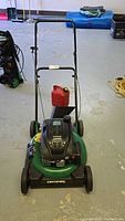 Front view of the Certified 21-inch gas push lawn mower with the gas can placed behind it on the floor. The mower has a black handle and green deck.