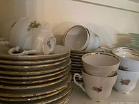 Stacked white Limoges china plates with floral design and gold edge, cups and saucers stacked nearby