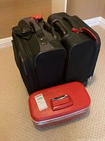 Front view of two black soft-sided suitcases and one red hard sided makeup case on carpeted floor.