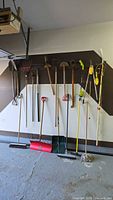 Photo showing a variety of long-handled garden and yard tools including rakes, shovels, hoes, a mop, and powered garden tools mounted on wall hooks.