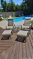 Full view of 2 beige mesh reclining chairs, 2 ottomans, and round side table arranged beside pool on wooden deck