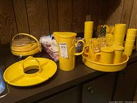 Yellow plastic picnic ware on a counter including serving trays, stack of plates, cups, pitcher, and utensil holder.