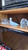 Shelf displaying two clear art glass sculptures with Viking ship designs and one gray stone bird sculpture between them, all placed on a wooden shelf.