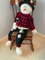 Front full view of plush snowman seated on wooden chair showing facial details, clothing, and small snowman plush in hand.