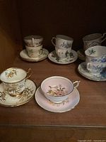 Overview of six different antique teacups and saucers with floral and gold designs arranged on wooden shelf.