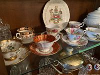 Display of assorted vintage teacups and saucers on glass shelf, showing floral patterns and gold trim. Canada decorative plate and tureen included.
