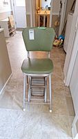 Front view of vinyl and metal vintage step stool with olive green padded seat and backrest showing scratches and wear.