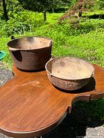 Two cast iron pots on a wooden surface outdoors, showing exterior surfaces heavily rusted and one pot larger with partial marking.