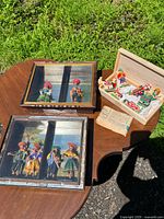 Photo showing two folk art shadow boxes with miniature dolls and an open wooden box with additional dolls on a table outdoors