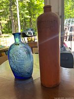 Photo of cobalt blue glass flask with embossed eagle and shield beside a tall brown stoneware bottle