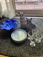 Photo showing all items: blue glass bowl, ceramic cats, stacked plates, and four glass goblets on carpet near window
