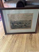 Framed full artwork depicting a cavalry charge battle scene, positioned on the floor with couch in background. Wooden frame, glass front, light tan mat.
