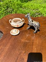Photo of ceramic covered dish with matching lid, floral decoration, next to ceramic zebra figurine on wooden table outdoors.