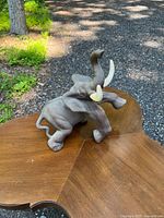 Elephant figurine shown from a side angle, placed on a wooden table outdoors with shaded ground and greenery in the background.