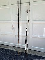 Full view of two fishing rods leaning against white garage door, showing rod length, handle, and line guides.