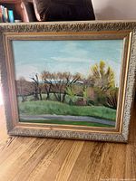 Full view of framed square landscape painting showing trees and a grassy area under a blue sky. Oval gilded frame with elaborate carving.