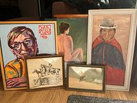 Set of 5 framed paintings arranged on floor showing colorful portrait, figurative nude, figure in bowler hat, and two smaller framed artworks (dog in snow, horse and rider sketch)