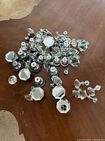 Top-down view of about two dozen assorted glass or crystal drawer pulls and knobs with attached metal hardware laying scattered on a wooden surface.