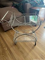 Full view of gold metal bar cart showing curved frame, two clear glass shelves, scroll design on handle, and caster wheels on wood floor.