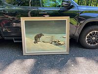 Photo showing full front view of large framed painting propped against a black vehicle outdoors. The artwork depicts a wolf overlooking a snowy village under a crescent moon sky, with wood frame visible.
