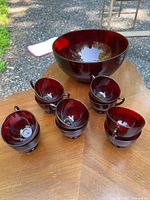 Overall view of the ruby red glass punch bowl with five of the matching cups arranged around it on a wooden table outdoors.
