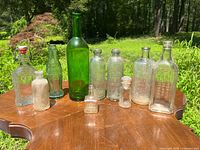 Lot of 10 vintage and antique glass bottles displayed outdoors in sunlight on wooden surface showing various shapes and styles including green and clear glass bottles.