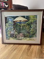 Framed watercolor painting of a garden with a white umbrella and various greenery, showing wood frame and cream-colored mat.