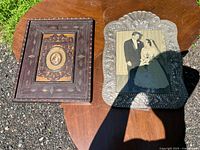 Two vintage picture frames placed on a wooden surface outdoors: On the right, an ornate metal frame embossed with floral and shell patterns containing a black and white wedding photo; on the left, a dark wooden frame with inlaid details holding a small floral oval insert.