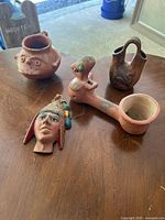 Four Pre-Columbian-inspired ceramic/clay pottery pieces shown on wooden surface: round face vessel, two-spouted handle vessel, bird-shaped pipe/ladle, and decorative head ornament with headdress.