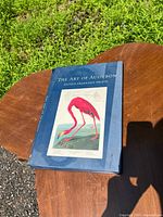 Blue hardcover book titled 'The Art of Audubon' featuring a flamingo bird illustration on the cover, placed on a wooden table outside.
