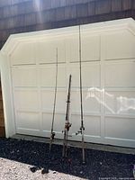 Three fishing rods standing upright outdoors against a garage door, showing length and overall appearance.