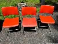 Three vintage Mid-Century Modern style chairs with orange plastic seats and backs, metal tubular frames shown outdoors on pavement.