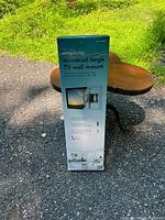 Box of Simplicity SXDP3-B1 universal large TV wall mount shown standing upright on outdoor surface.
