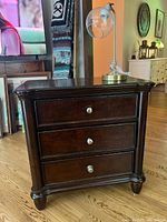 Full view of small dark brown wooden chest of drawers with metal knobs and table lamp on top, shown in living room setting.