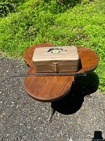 Photo of a brown vintage metal storage box with a National Rifle Association emblem on lid, two metal cleaning rods resting on a wooden table with green grass background.