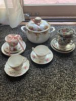 Full lot showing seven teacups and seven saucers with floral design and white porcelain lidded pot with floral and gold decoration placed on a carpeted surface near window.