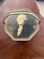 Top view of octagonal biscuit tin lid with George Washington portrait, showing wear and scratches.