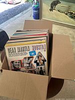 Box filled with multiple 12-inch vinyl record LPs. The most visible album is Dean Martin Houston with colorful cover and photographic elements.
