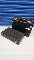 Two black cases; one larger briefcase-style with metal latch lock and key lock, one smaller board game case with two gold latch locks.