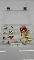 Clear plastic tackle box open showing assorted fishing lures including soft baits and hard lures in multiple compartments, alongside packaged soft bait.