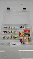 Clear plastic fishing tackle box open showing multiple compartments filled with various fishing lures and accessories including spoons, jigheads, and packaged terminal tackle.