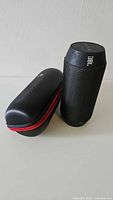 Black cylindrical JBL Pulse speaker with mesh grille, standing upright next to black semi-hard carrying case with red zipper.