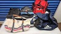 Overall view of 5 bags on table showing different colors, sizes and logos from Adidas, Atomic and Hilfiger brands.