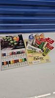 Overview image showing all items including closed oil colour box with cover art alongside acrylic paint tubes, bottles, and palette pad.