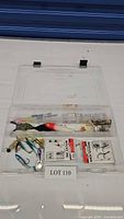 Clear plastic fishing tackle box opened showing assorted fishing lures, hooks, and spinner baits inside with visible rust in some compartments