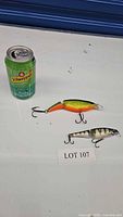 Two fishing lures displayed with a soda can for size reference. One lure is segmented with bright green/yellow/orange colors, the other is silver with green and black stripes.