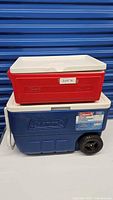 Stacked two Coleman coolers, small red cooler on top of larger blue wheeled cooler with white lids.