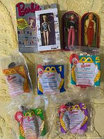 Lot contents showing 2 boxed Barbie dolls and 5 McDonald's Barbie Happy Meal toys in original sealed plastic packages on a yellow surface.